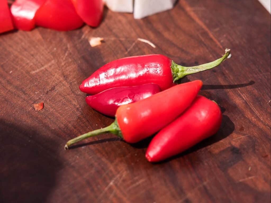 BEST Baby Bok Choy Kimchi Recipe - Easy & Delicious 15 Three red chili peppers with green stems rest on a dark wooden surface. In the background, there are a few chopped red and white vegetables slightly out of focus, hinting at the vibrant ingredients often found in bok choy kimchi.