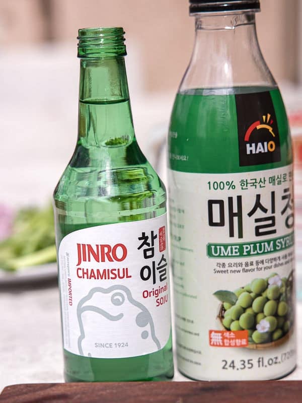 BEST Baby Bok Choy Kimchi Recipe - Easy & Delicious 11 A green bottle of Jinro Chamisul soju and a larger bottle of Haio ume plum syrup sit side by side on a table, accompanied by a small dish of bok choy kimchi, with blurred food items in the background.