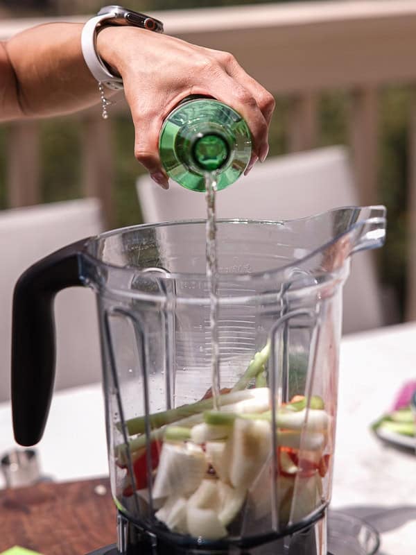 BEST Baby Bok Choy Kimchi Recipe - Easy & Delicious 33 A hand pours clear liquid from a green bottle into a blender filled with chopped vegetables like onions, celery, and bok choy kimchi, outdoors on a sunny day.