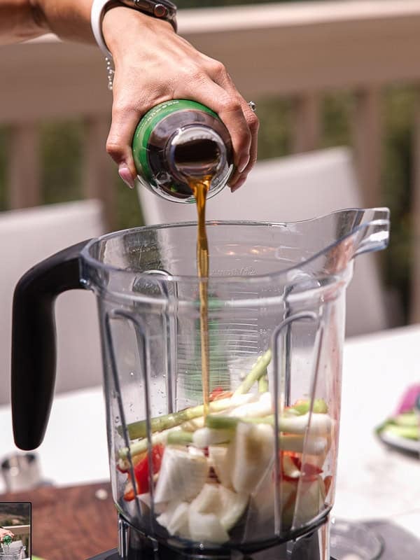 BEST Baby Bok Choy Kimchi Recipe - Easy & Delicious 32 A hand pours liquid from a bottle into a blender filled with cut vegetables, including onions, asparagus, and bok choy kimchi, outdoors on a sunny day.