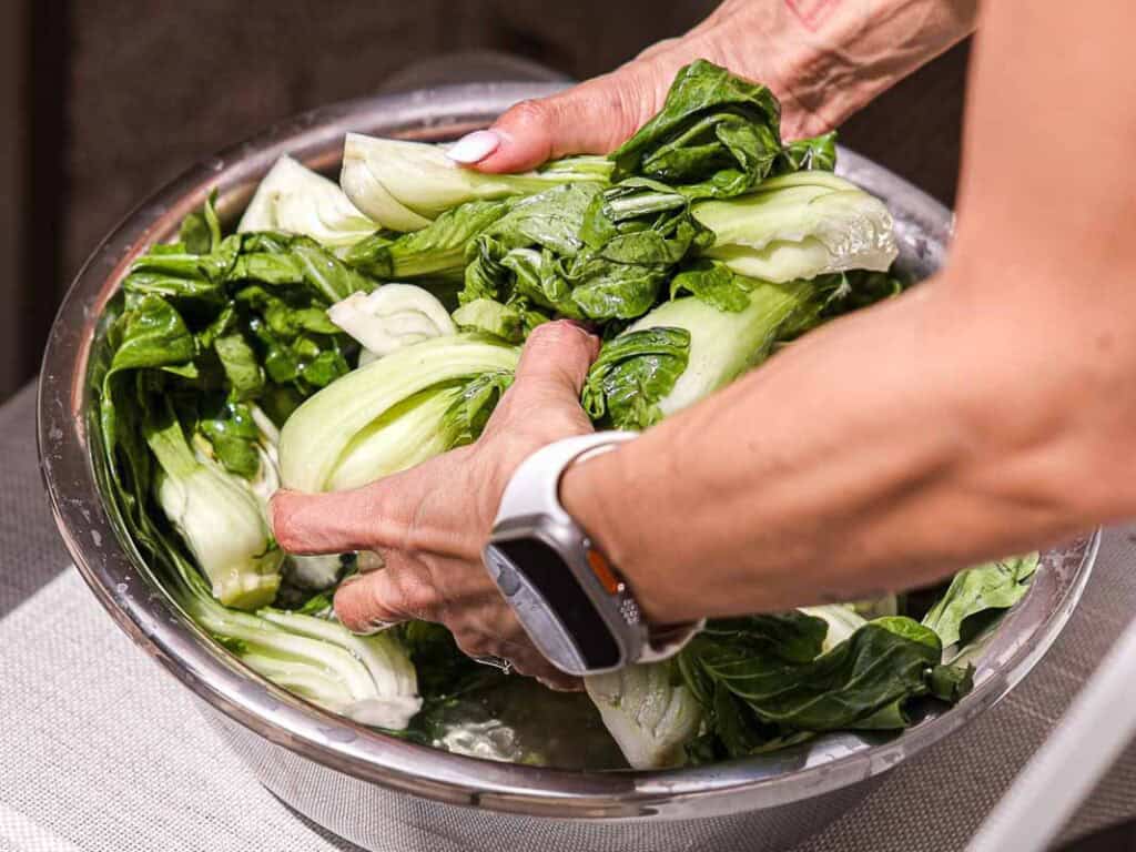 BEST Baby Bok Choy Kimchi Recipe - Easy & Delicious 27 A person wearing a white wristwatch washes fresh bok choy in a metal bowl filled with water, preparing the vegetables for homemade bok choy kimchi.