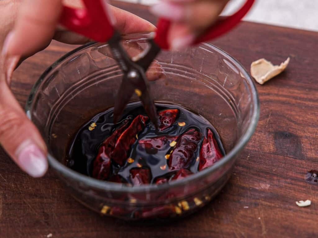 BEST Baby Bok Choy Kimchi Recipe - Easy & Delicious 30 Hands use red scissors to cut dried red chiles in a glass bowl filled with dark soy sauce, all on a wooden surface. Some chile seeds and a garlic peel are visible nearby, alongside a small portion of bok choy kimchi tucked to the side.