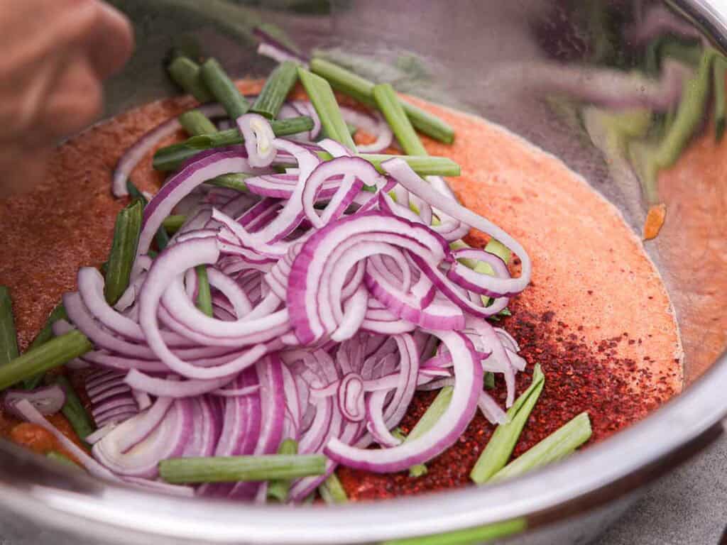 BEST Baby Bok Choy Kimchi Recipe - Easy & Delicious 40 A metal bowl containing sliced red onions, green beans, bok choy kimchi, red chili flakes, and reddish marinade, with a hand stirring the mixture.