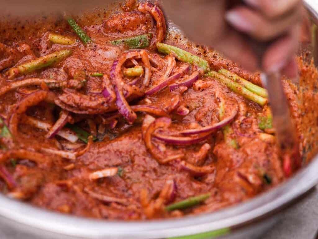 BEST Baby Bok Choy Kimchi Recipe - Easy & Delicious 41 A close-up of a bowl containing sliced red onions and green vegetables, including bok choy kimchi, mixed with a red, spicy marinade, being stirred by a personโs hand.