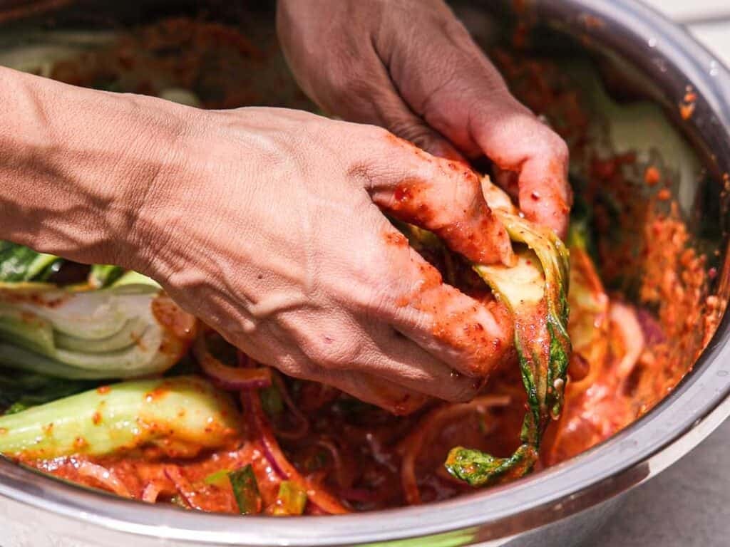 BEST Baby Bok Choy Kimchi Recipe - Easy & Delicious 44 Hands mixing leafy vegetables with red seasoning in a metal bowl, preparing bok choy kimchi.