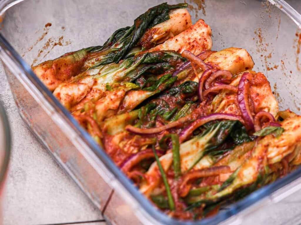 BEST Baby Bok Choy Kimchi Recipe - Easy & Delicious 2 Glass container filled with homemade kimchi, featuring napa cabbage, bok choy kimchi, green onions, and sliced red onion coated in a red chili pepper seasoning paste.