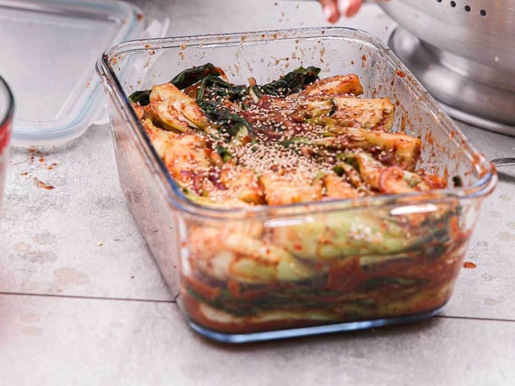 BEST Baby Bok Choy Kimchi Recipe - Easy & Delicious 46 A glass container filled with homemade bok choy kimchi, garnished with sesame seeds, sits on a gray countertop. Some leafy greens are visible among the red, spicy cabbage.