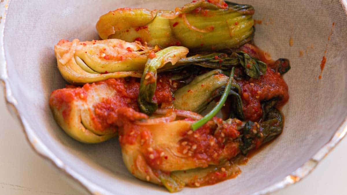 BEST Baby Bok Choy Kimchi Recipe - Easy & Delicious 1 A bowl of bok choy kimchi covered in red chili sauce, served in a grayish ceramic bowl on a white surface.