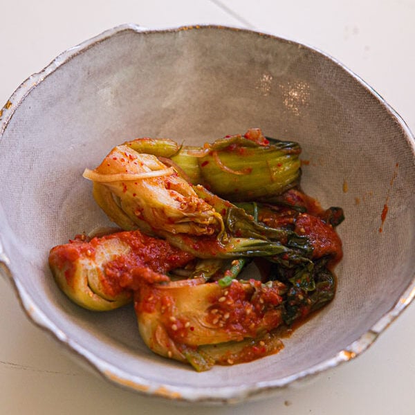 A ceramic bowl containing bok choy kimchi covered in a red, spicy sauce sits on a white surface.